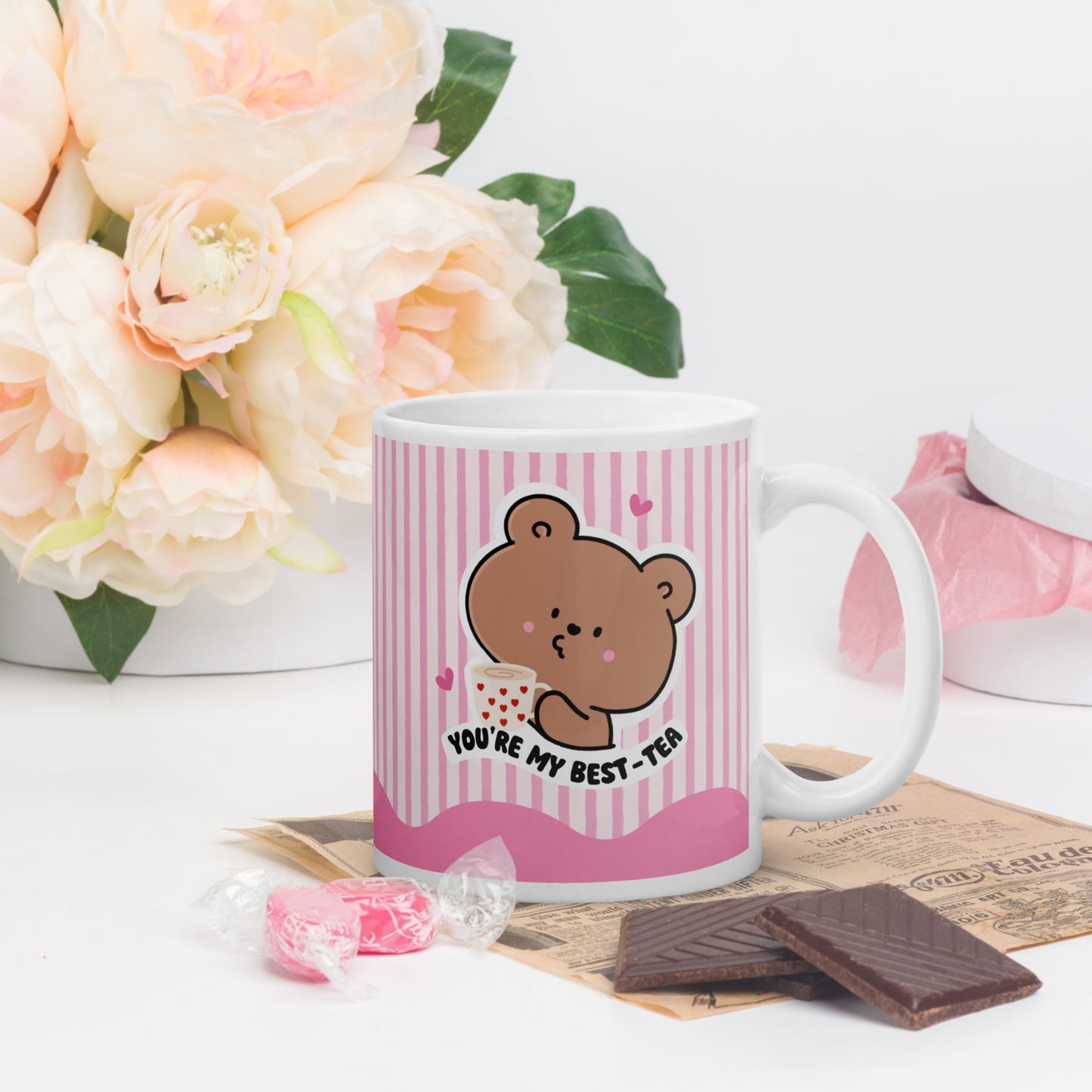 Mug with a bear design and text, surrounded by flowers and snacks on a white surface