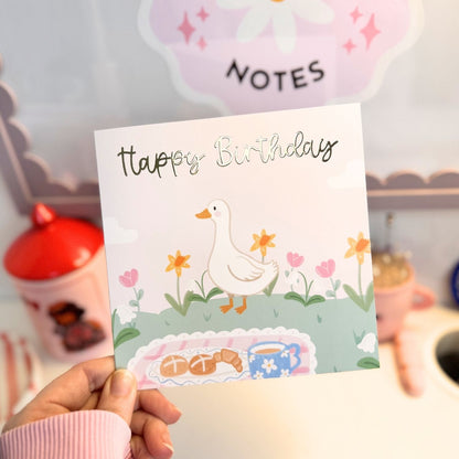 A hand holds a square birthday card featuring a cute illustration of a white duck walking among flowers, with the words 'Happy Birthday' written above. The background is a softly blurred pastel room with a pink 'NOTES' sign and decorative objects.