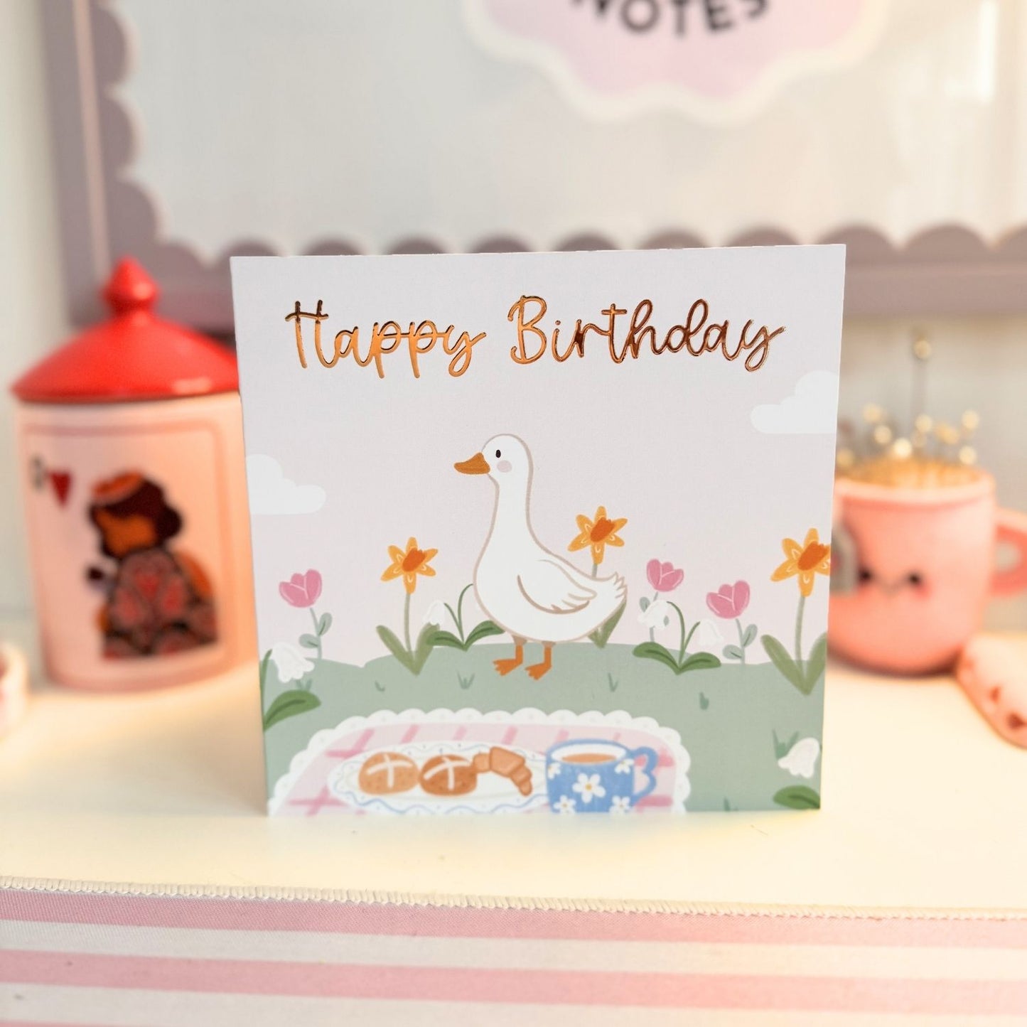A square birthday card with a white background featuring an illustration of a white duck walking among flowers, with 'Happy Birthday' written in metallic gold script. The card is displayed standing upright on a shelf with pink decorative items blurred in the background.