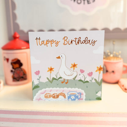 A square birthday card with a white background featuring an illustration of a white duck walking among flowers, with 'Happy Birthday' written in metallic gold script. The card is displayed standing upright on a shelf with pink decorative items blurred in the background.