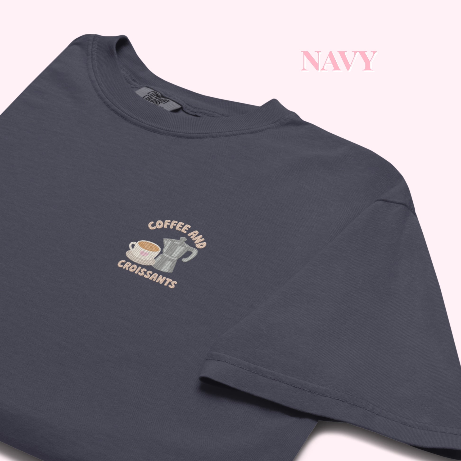 close up Embroidered navy tshirt with coffee and croissant text and a cute coffee mug and grey stove top coffee maker. slow cozy moments. coffee lover gift