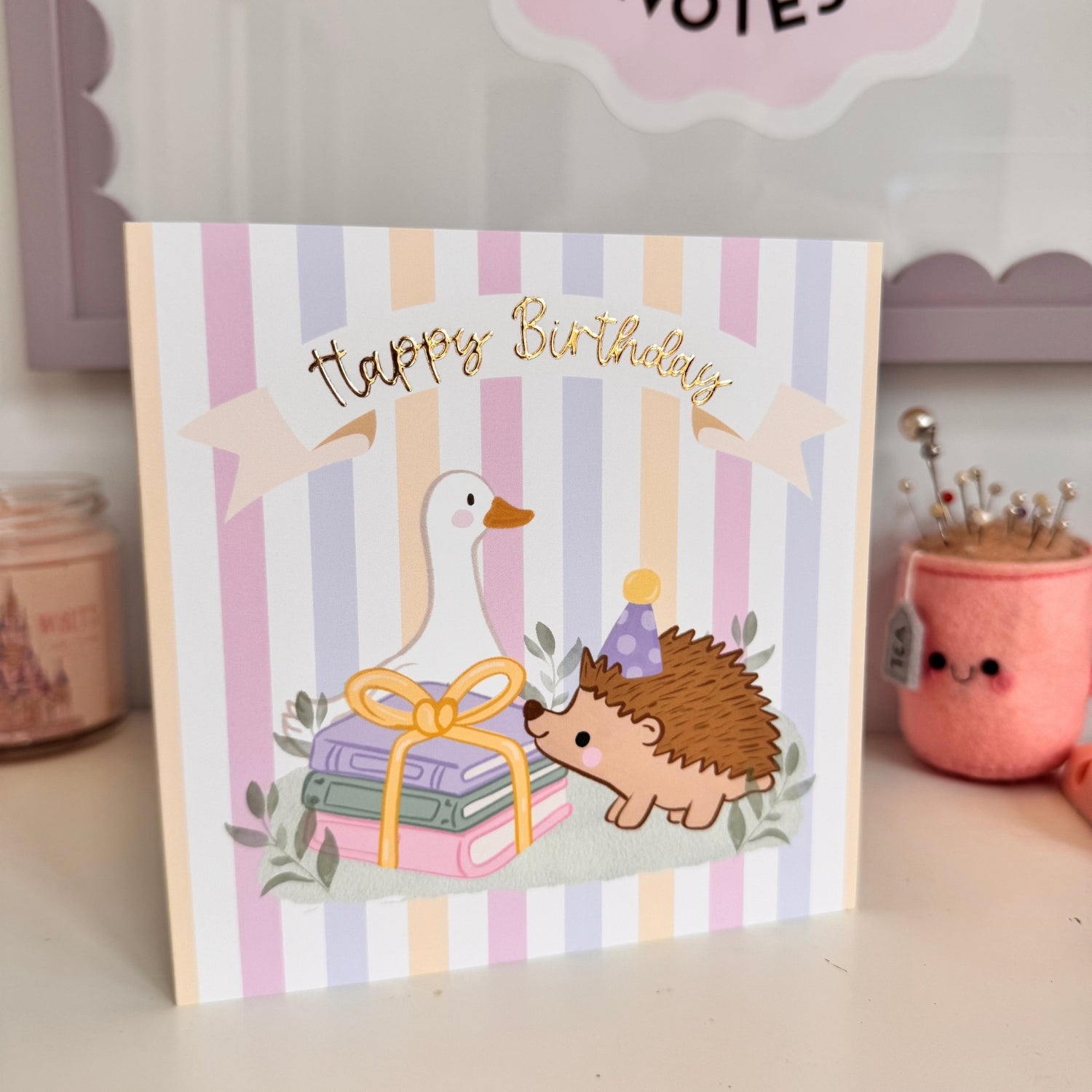 A square birthday card with pastel vertical stripes and a cute illustration of a white goose, a small hedgehog wearing a party hat, and stacked gift boxes under a gold 'Happy Birthday' banner. The card is photographed standing on a tabletop with soft-focus decorative objects in the background.