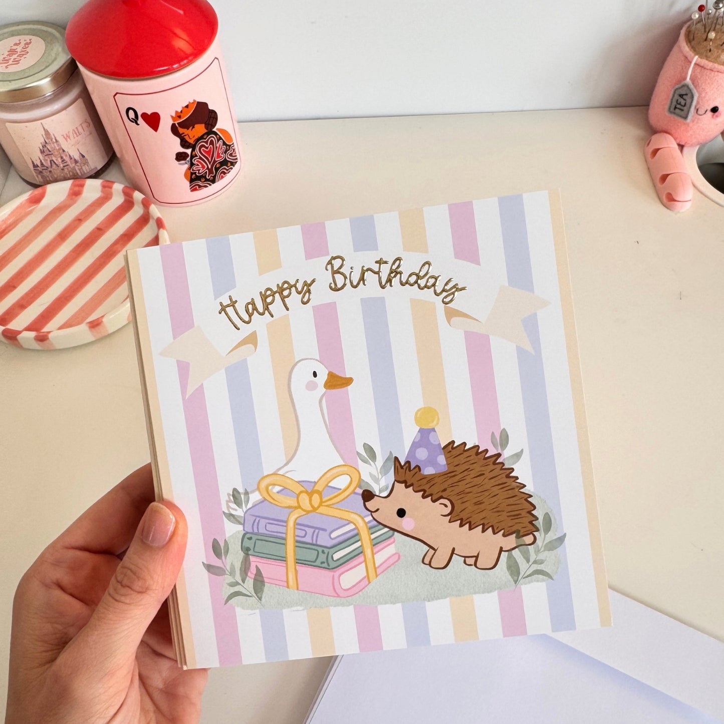 A hand holds a pastel striped 'Happy Birthday' greeting card featuring a white goose and a hedgehog illustration. The card is shown in a cozy tabletop setting with other decorative items in the background.