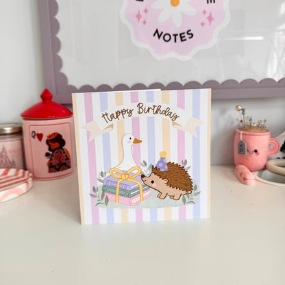 A square birthday card with pastel vertical stripes and a cute illustration of a white goose, a hedgehog in a party hat, and stacked presents, with the words 'Happy Birthday' across the top. The card is displayed on a white tabletop with pink decorative items in the background.