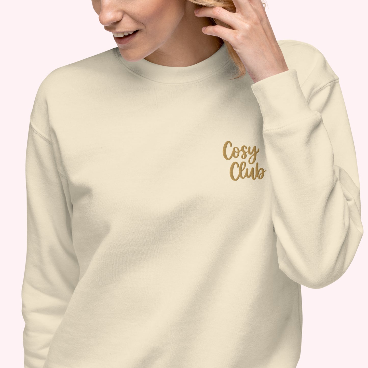 A woman wearing a cream-colored crewneck sweatshirt with 'Cosy Club' embroidered in gold script on the left chest, set against a solid pale pink background.