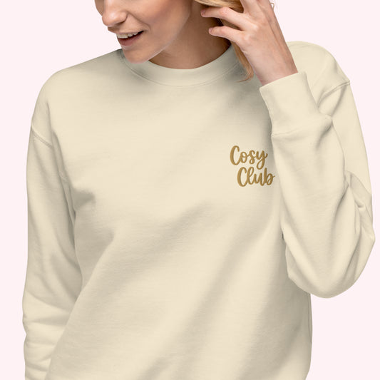 A woman wearing a cream-colored crewneck sweatshirt with 'Cosy Club' embroidered in gold script on the left chest, set against a solid pale pink background.