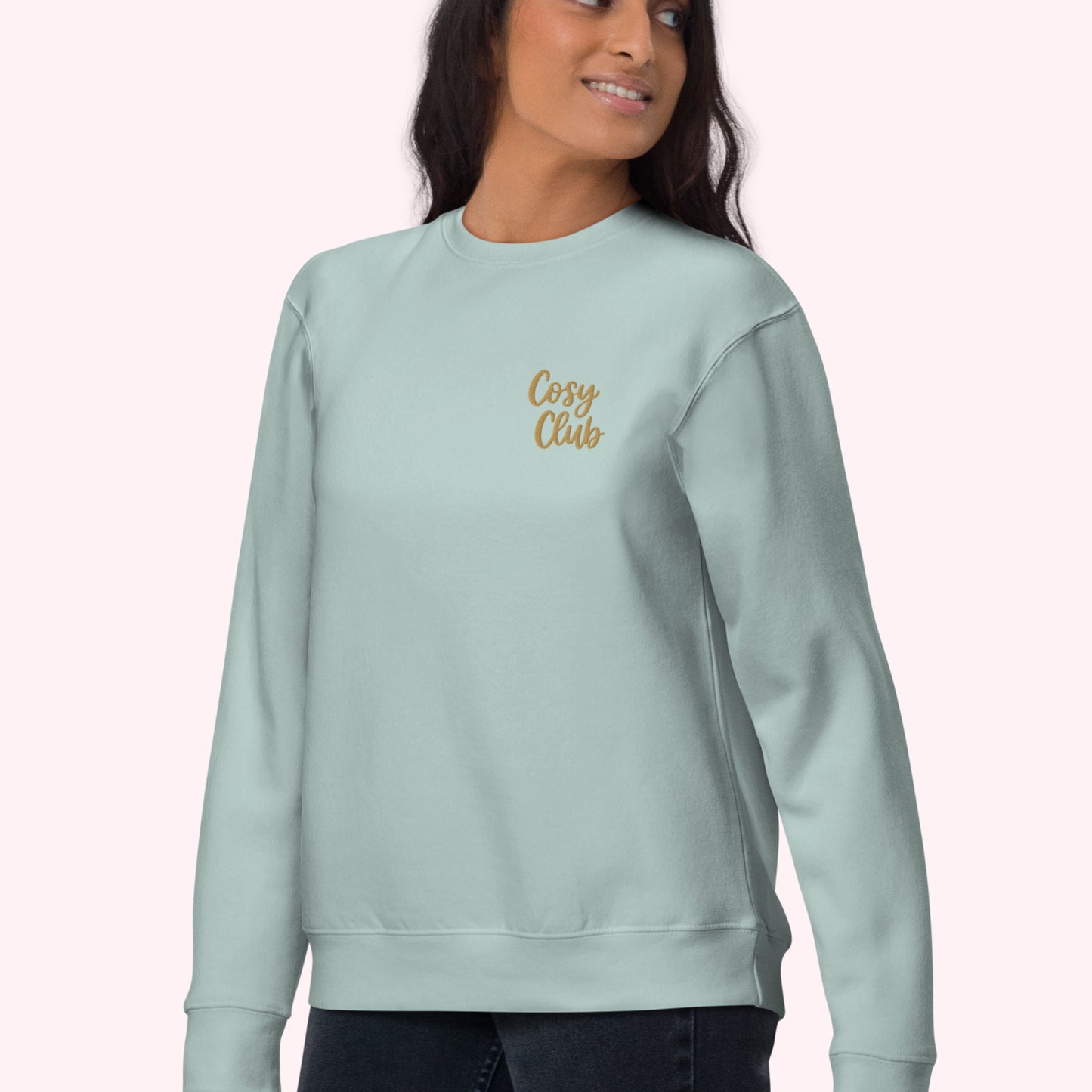 A woman wearing a light mint green crewneck sweatshirt with 'Cosy Club' embroidered in gold script on the left chest, set against a solid pale pink background.