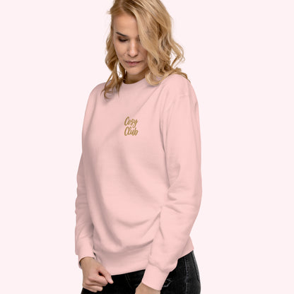 A woman wearing a light pink crewneck sweatshirt with 'Cozy Club' embroidered in gold script on the left chest, set against a solid pale pink background.
