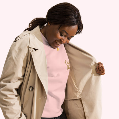 A woman wearing a light beige trench coat over a light pink crewneck sweatshirt with gold embroidered cosy club, posing against a solid pale pink background.