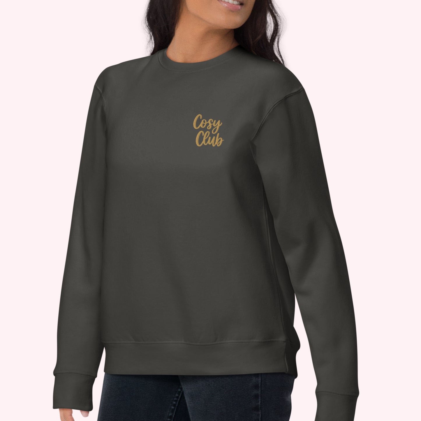 A woman wearing a dark charcoal gray crewneck sweatshirt with 'Cosy Club' embroidered in gold script on the left chest, paired with dark blue jeans against a pale pink background.