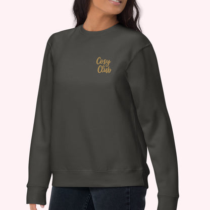 A woman wearing a dark charcoal gray crewneck sweatshirt with 'Cosy Club' embroidered in gold script on the left chest, paired with dark blue jeans against a pale pink background.