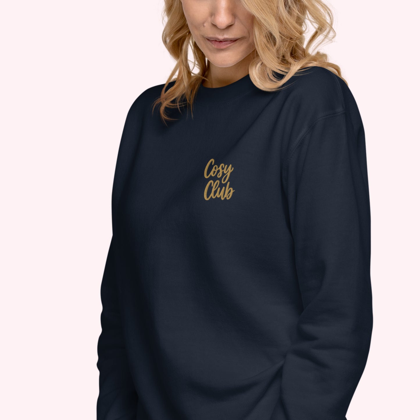 A woman wearing a dark navy crewneck sweatshirt with 'Cosy Club' embroidered in gold script on the left chest, set against a solid pale pink background.