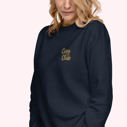 A woman wearing a dark navy crewneck sweatshirt with 'Cosy Club' embroidered in gold script on the left chest, set against a solid pale pink background.
