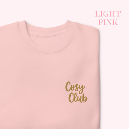 A folded light pink crewneck sweatshirt with 'Cosy Club' embroidered in gold script on the chest, shown against a pale pink background with 'LIGHT PINK' text in the upper right.