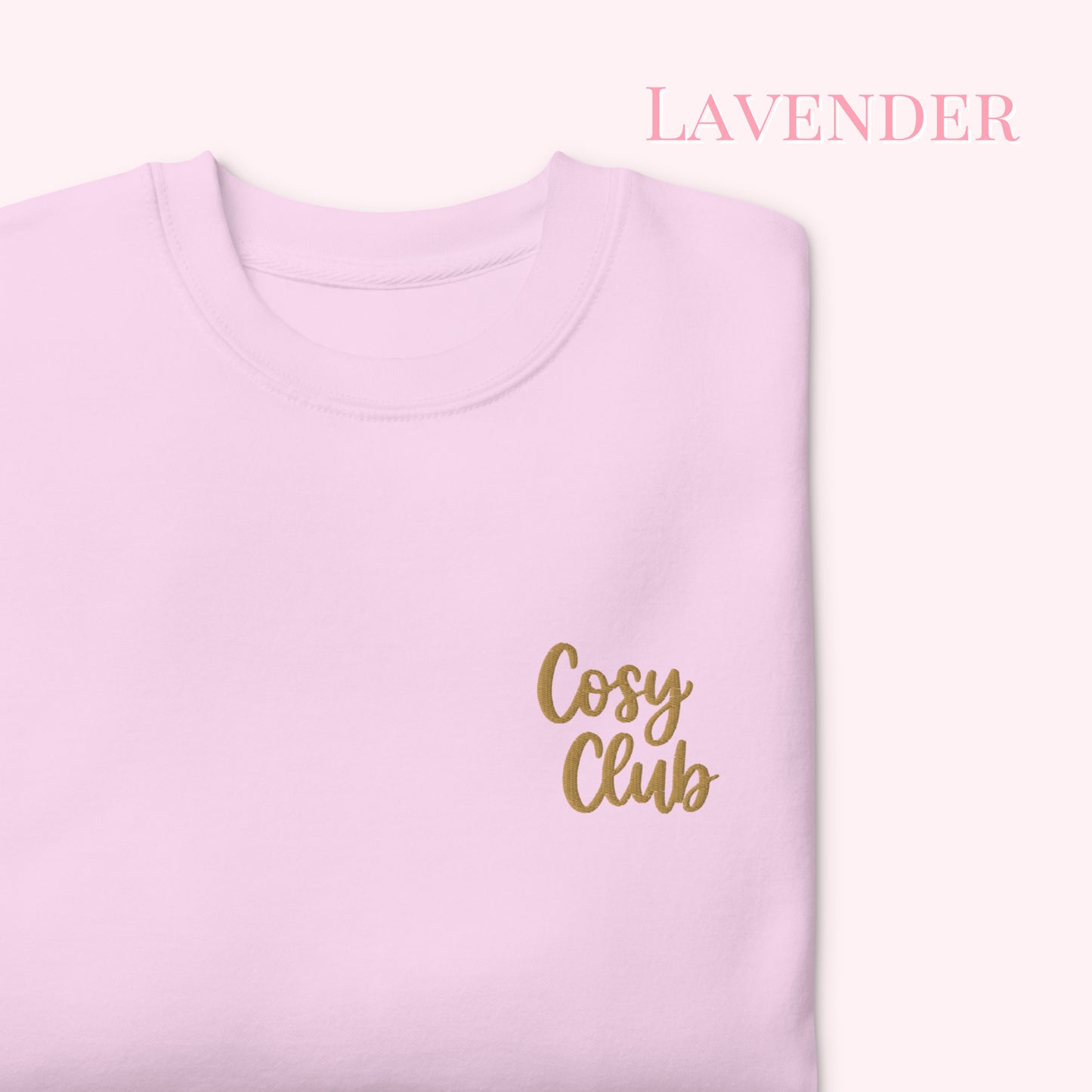 A folded light lavender crewneck sweatshirt with 'Cosy Club' embroidered in gold script on the chest, set against a pale pink background with 'LAVENDER' text in the upper right.