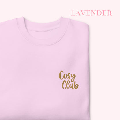 A folded light lavender crewneck sweatshirt with 'Cosy Club' embroidered in gold script on the chest, set against a pale pink background with 'LAVENDER' text in the upper right.
