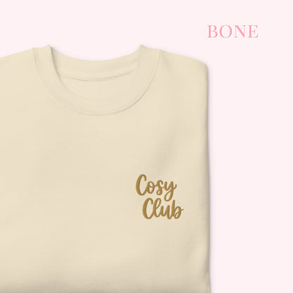 A folded cream-colored jumper with 'Cosy Club' embroidered in gold script on the chest, set against a pale pink background with the word 'BONE' in pink text in the upper right.