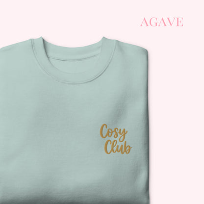 A folded sage green crewneck sweatshirt with 'Cosy Club' embroidered in gold script on the left chest, set against a pale pink background with the word 'AGAVE' in pink text in the upper right.