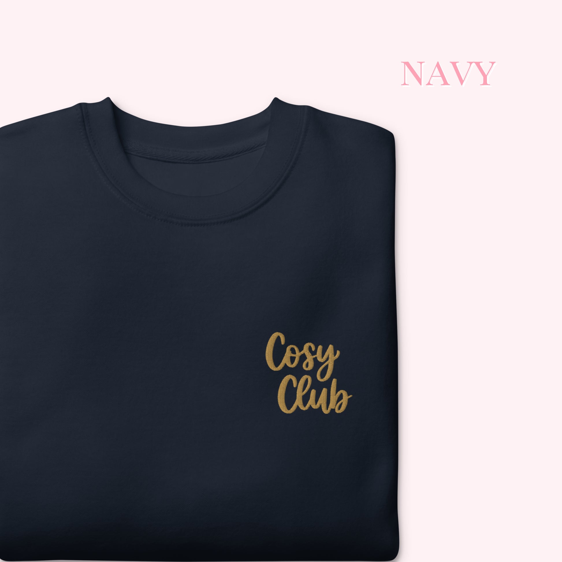 A folded navy crewneck sweatshirt with 'Cosy Club' embroidered in gold script on the chest, displayed on a pale pink background with the word 'NAVY' in the top right corner.
