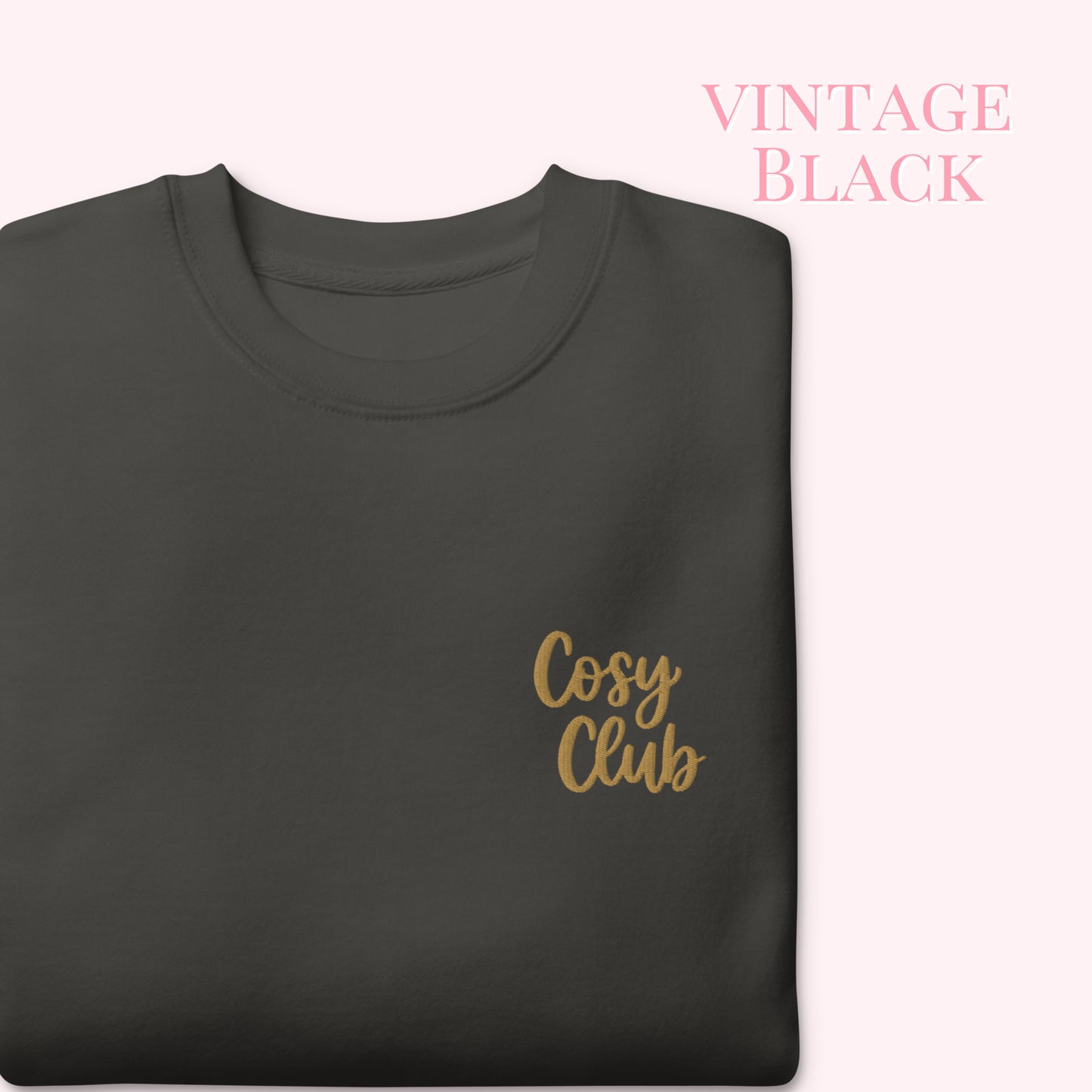 A folded dark charcoal gray t-shirt with 'Cosy Club' embroidered in gold script on the left chest, set against a pale pink background with 'VINTAGE BLACK' text in the upper right.