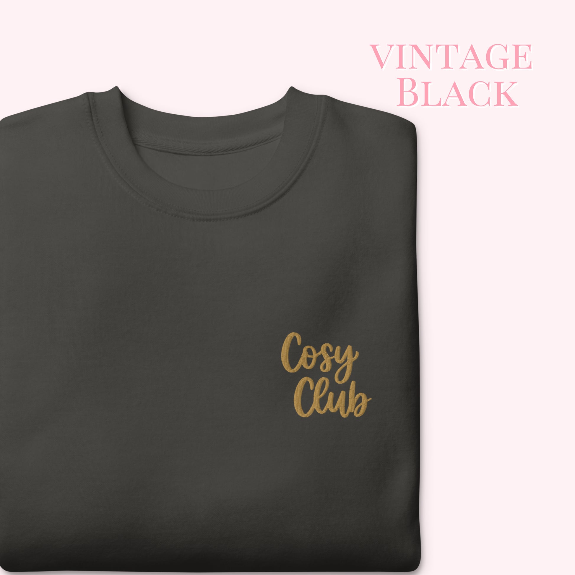 A folded dark charcoal gray t-shirt with 'Cosy Club' embroidered in gold script on the left chest, set against a pale pink background with 'VINTAGE BLACK' text in the upper right.
