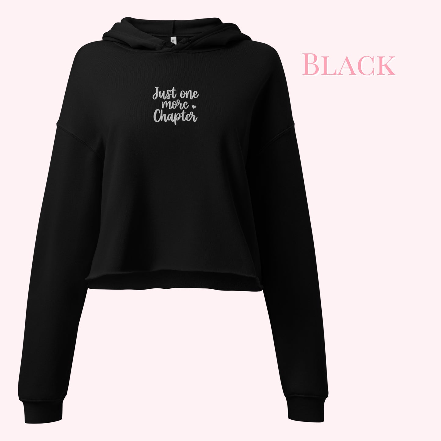 A black cropped hoodie sweatshirt with 'Just one more Chapter' embroidered in white script on the chest, displayed against a pale pink background with the word 'BLACK' in pink text in the upper right.