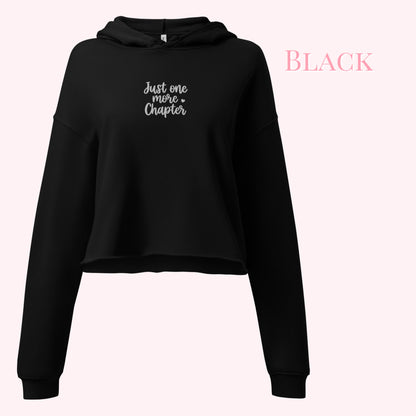 A black cropped hoodie sweatshirt with 'Just one more Chapter' embroidered in white script on the chest, displayed against a pale pink background with the word 'BLACK' in pink text in the upper right.