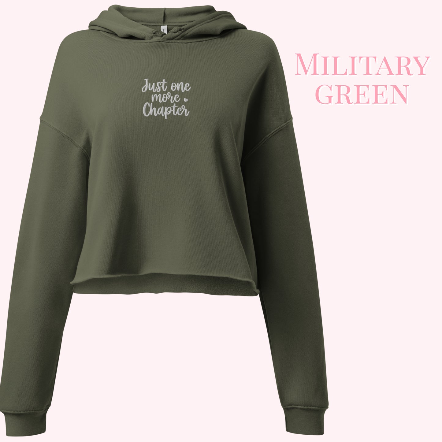 An olive green cropped hoodie with 'Just one more chapter' embroidered in white script on the chest, displayed against a pale pink background with 'MILITARY GREEN' text on the right.