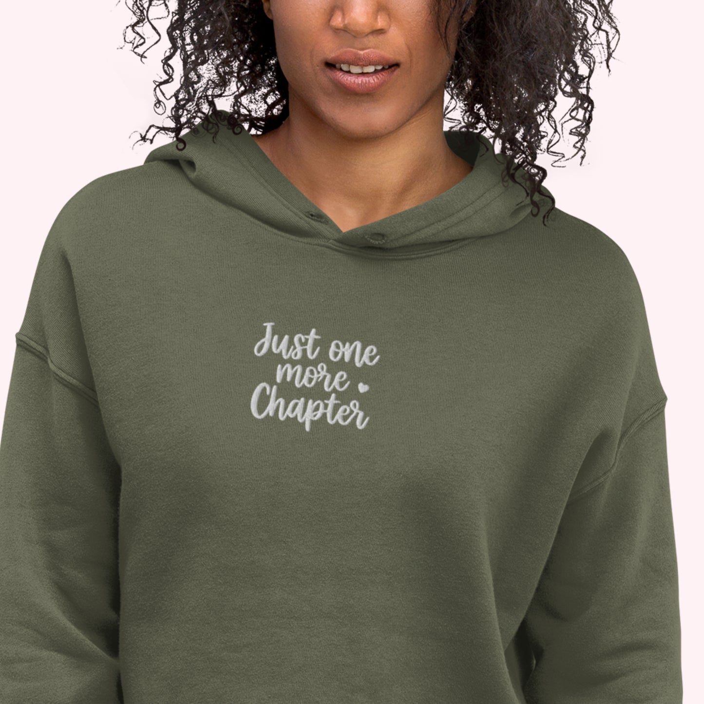 A woman wearing an olive green pullover hoodie with the text 'Just one more Chapter' printed in white script on the chest, set against a plain light pink background.