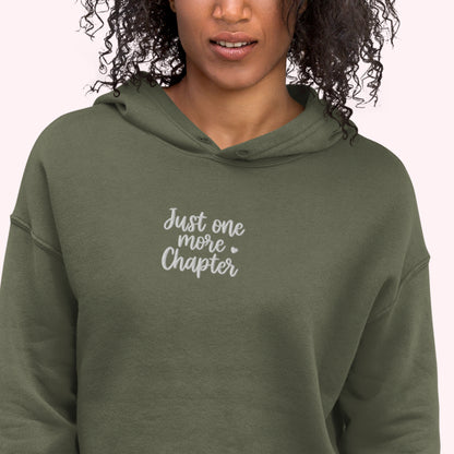 A woman wearing an olive green pullover hoodie with the text 'Just one more Chapter' printed in white script on the chest, set against a plain light pink background.