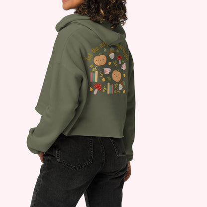 A woman wearing an olive green cropped hoodie with a colorful autumn bookish graphic print on the back, paired with black jeans, shown from a rear three-quarter angle against a light pink background.