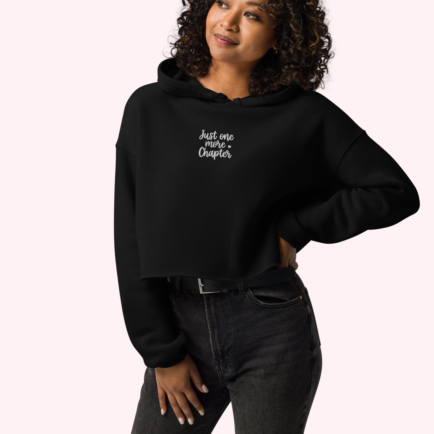 A woman wearing a black cropped hoodie with the white text 'Just one more chapter' on the chest, paired with dark gray jeans, against a solid pale pink background.