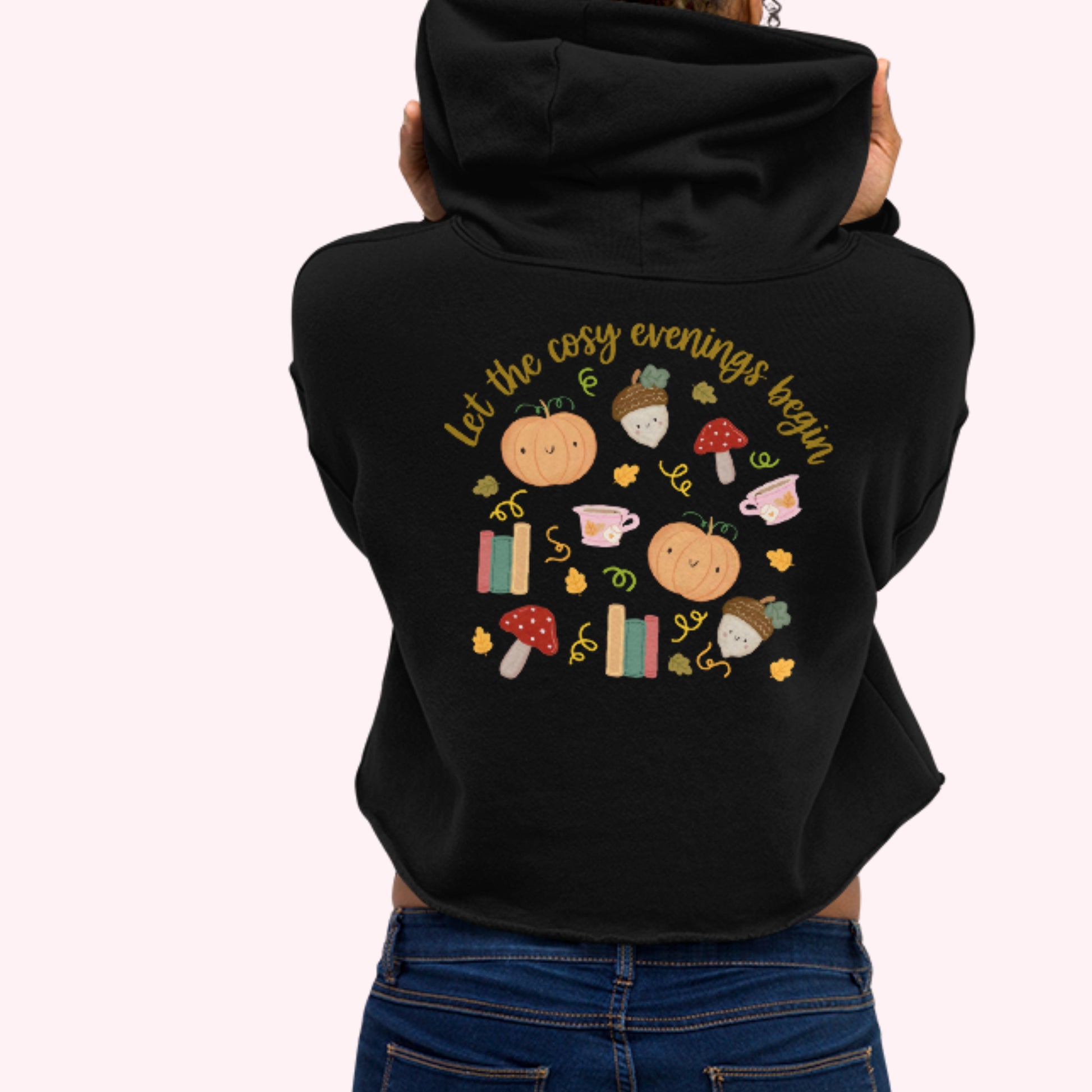 A women shown from the back wearing a black cropped hoodie with a colorful autumn-themed graphic print on the back, paired with blue jeans, against a light pink background. let the cosy evening begin logo