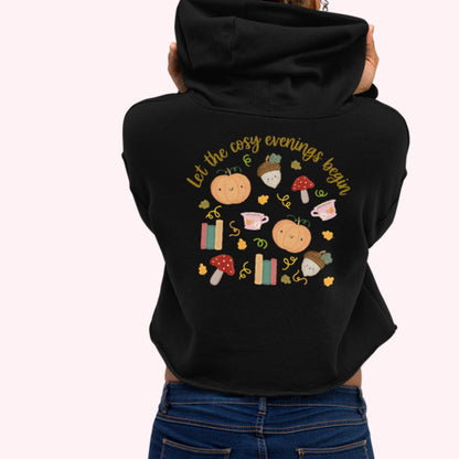 A women shown from the back wearing a black cropped hoodie with a colorful autumn-themed graphic print on the back, paired with blue jeans, against a light pink background. let the cosy evening begin logo