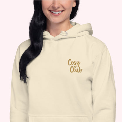 A woman wearing a cream-colored hoodie with 'Cosy Club' embroidered in gold script on the left chest, shown against a pale pink background.