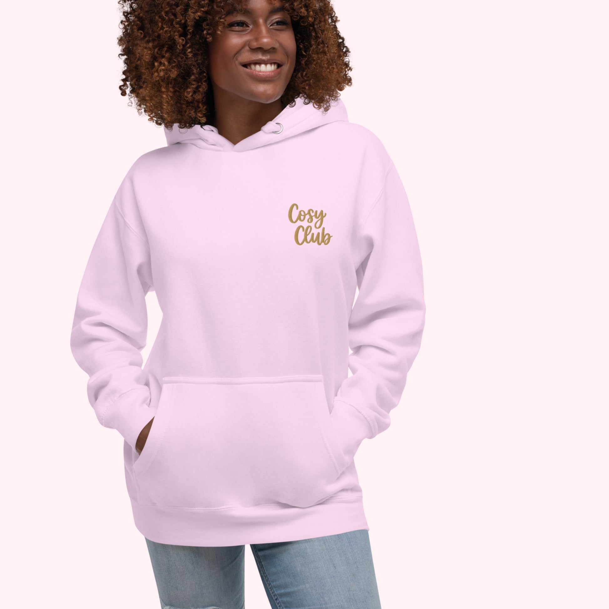 A smiling woman with curly brown hair wearing a light pink pullover hoodie with 'Cosy Club' embroidered in gold script on the left chest, paired with blue jeans, set against a solid pale pink background.