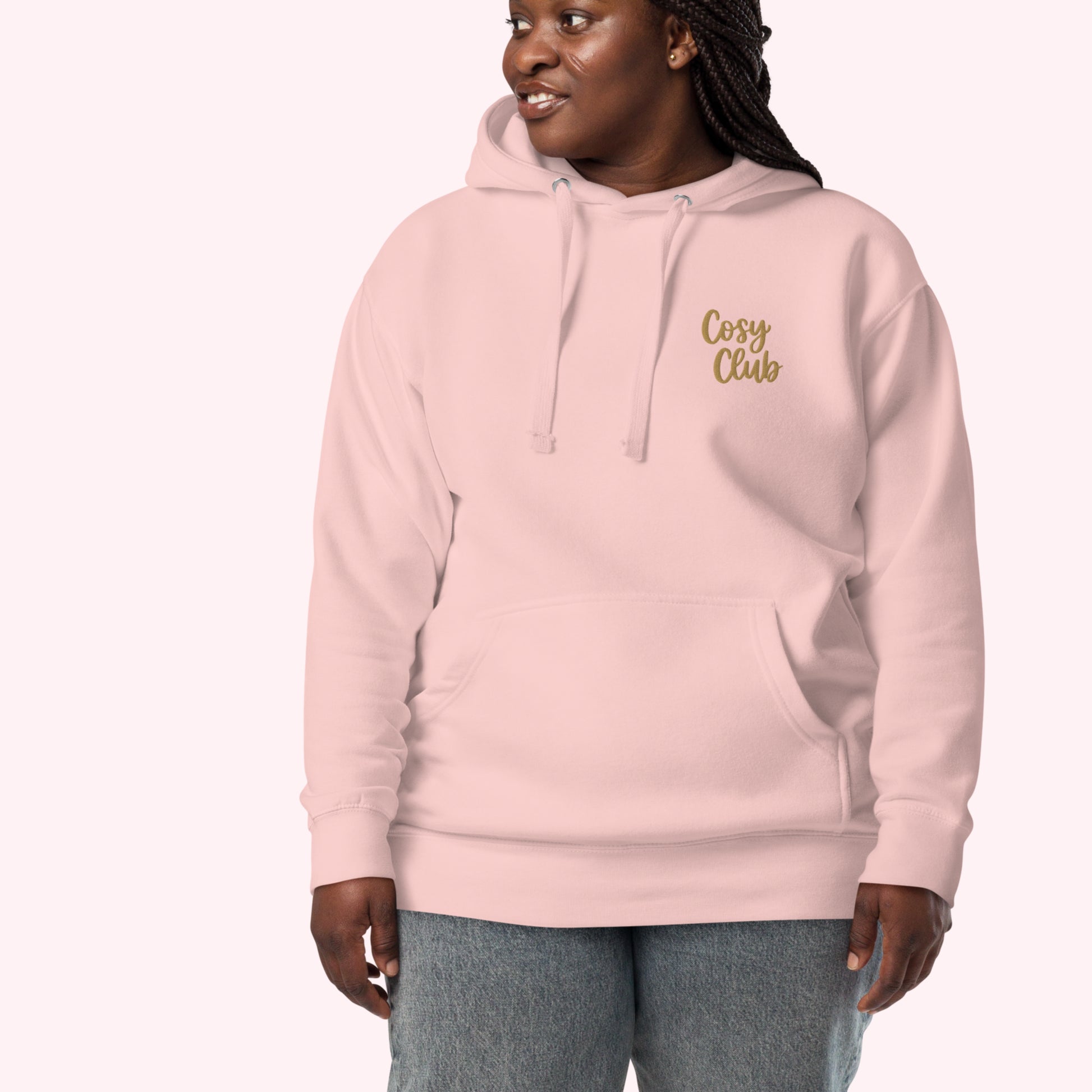 A woman wearing a light pink pullover hoodie with 'Cosy Club' embroidered in gold script on the left chest, paired with gray jeans, set against a solid pale pink background.