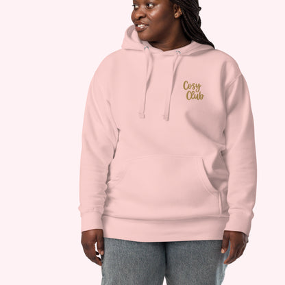 A woman wearing a light pink pullover hoodie with 'Cosy Club' embroidered in gold script on the left chest, paired with gray jeans, set against a solid pale pink background.