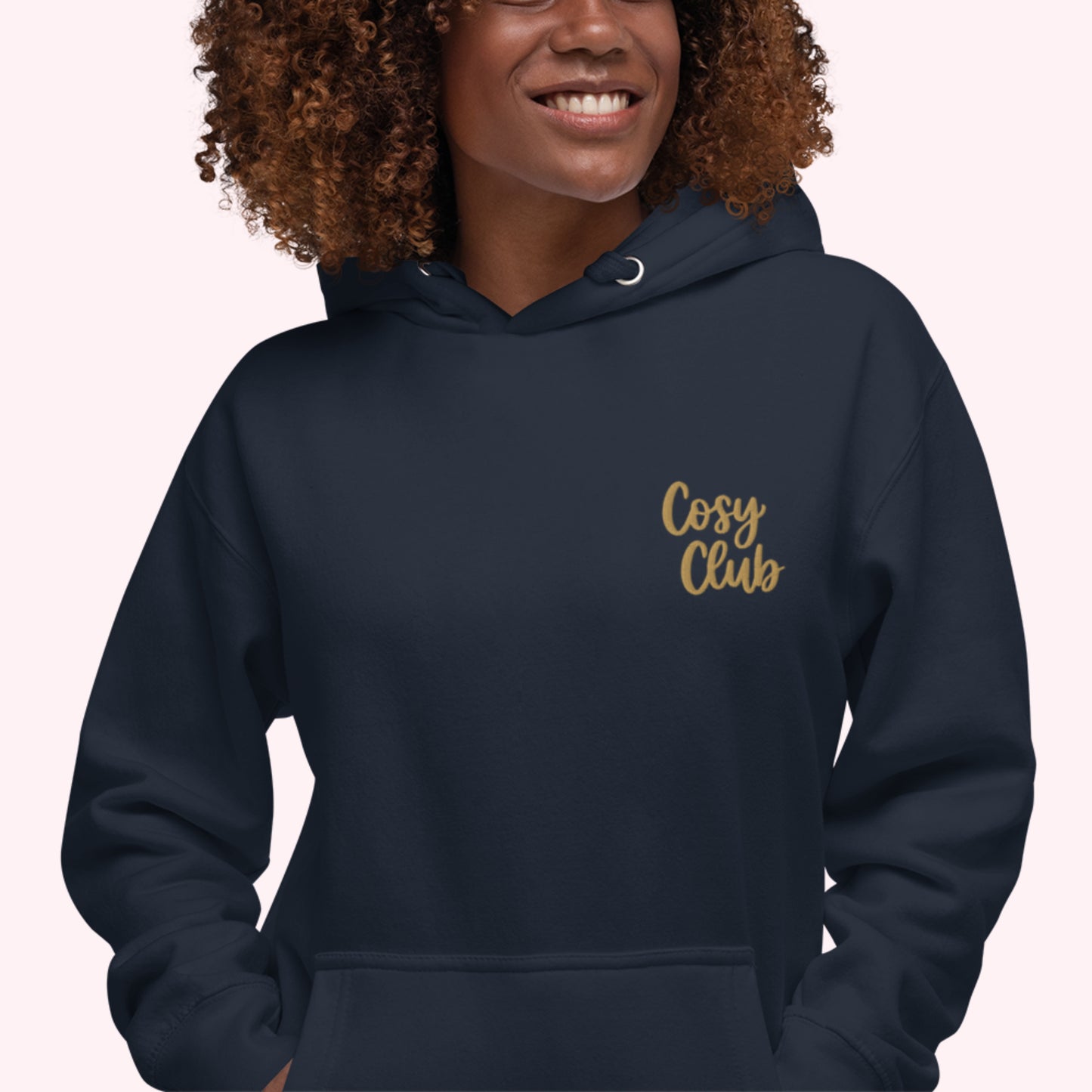 A woman wearing a navy blue hoodie with 'Cosy Club' embroidered in gold script on the left chest, set against a solid pale pink background.