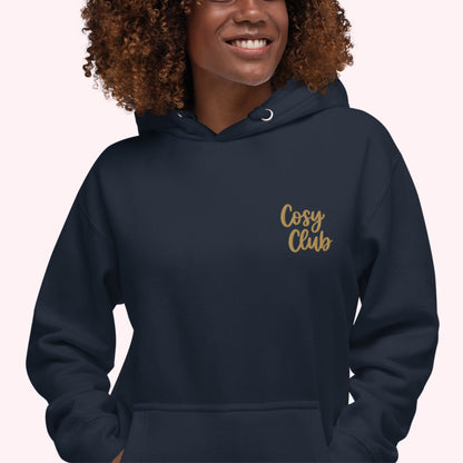 A woman wearing a navy blue hoodie with 'Cosy Club' embroidered in gold script on the left chest, set against a solid pale pink background.