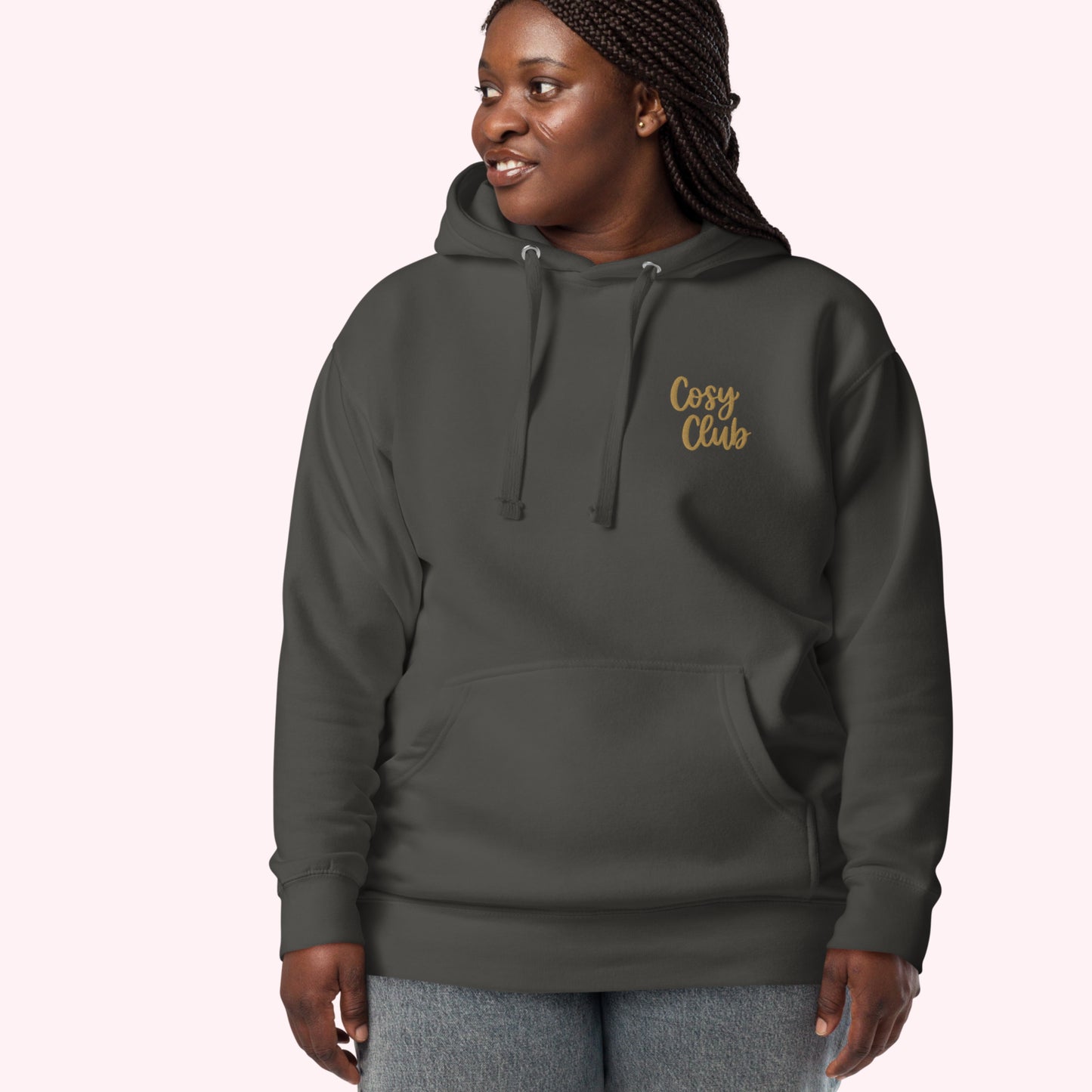 A woman wearing a dark charcoal gray hoodie with 'Cosy Club' embroidered in gold script on the left chest, paired with gray jeans, set against a solid pale pink background.