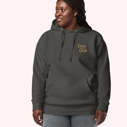 A woman wearing a dark charcoal gray hoodie with 'Cosy Club' embroidered in gold script on the left chest, paired with gray jeans, set against a solid pale pink background.