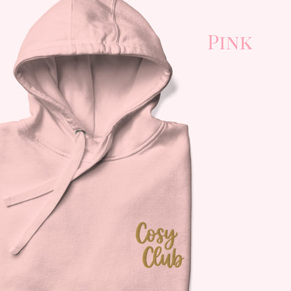 A folded light pink hoodie with a gold 'Cosy Club' logo on the front, displayed against a pale pink background with the word 'PINK' in the upper right.