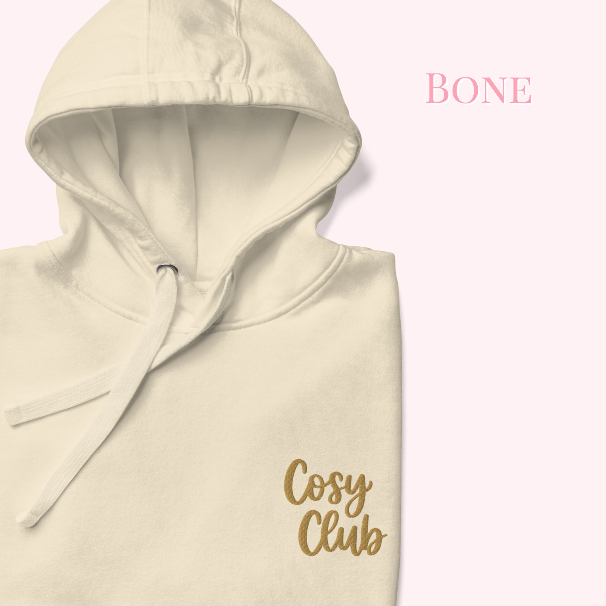 A cream-colored hoodie with a gold 'Cosy Club' logo on the chest, shown against a pale pink background with the text 'BONE' in the upper right.