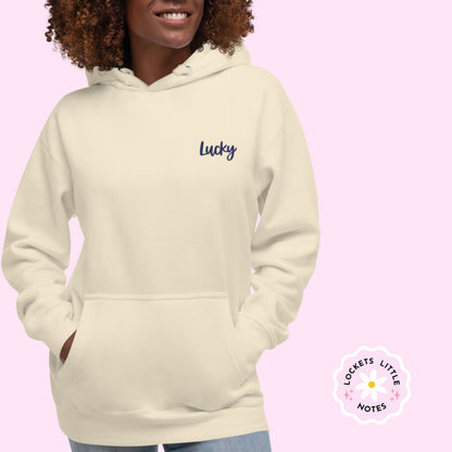 women wearing a beige hoodie with 'Lucky' text on a pink background and lockets little notes logo lucky charm hoody