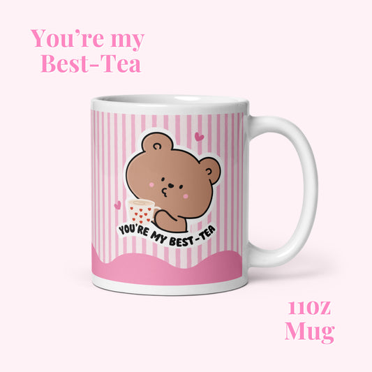 White glossy mug with a cute bear illustration design and text that says you're ny best-tea on a pink stripe background