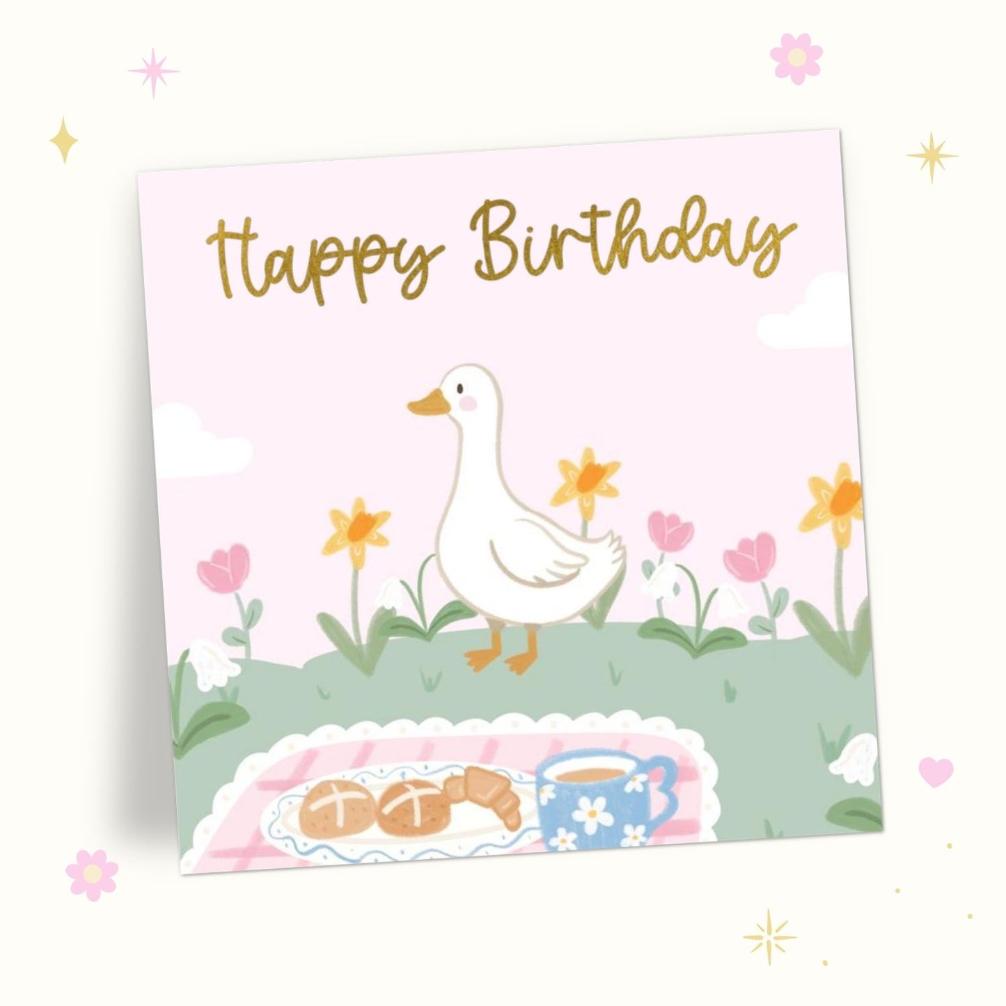 A square birthday card with a pastel illustration of a white duck walking among flowers, with the words 'Happy Birthday' in gold script at the top. A picnic blanket with pastries and a blue cup appears at the bottom.