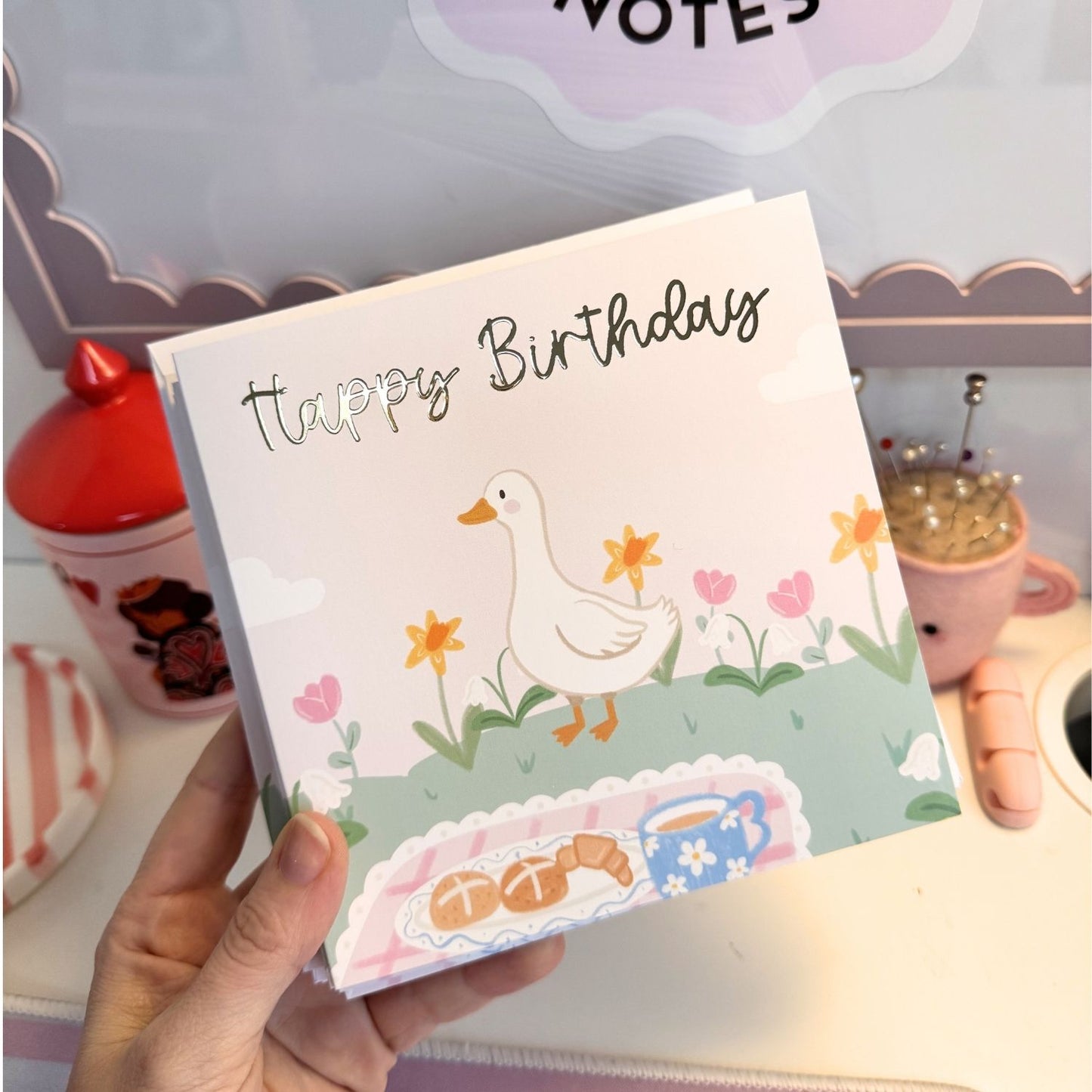 A hand holds a square greeting card with a pastel illustration of a white duck, flowers, and the words 'Happy Birthday' in black script. The card is photographed in a styled desk or craft room setting with a red container, a pin cushion, and other pastel objects in the background.
