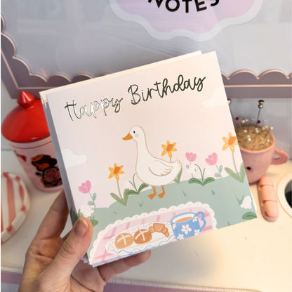 A hand holds a square greeting card with a pastel illustration of a white duck, flowers, and the words 'Happy Birthday' in black script. The card is photographed in a styled desk or craft room setting with a red container, a pin cushion, and other pastel objects in the background.