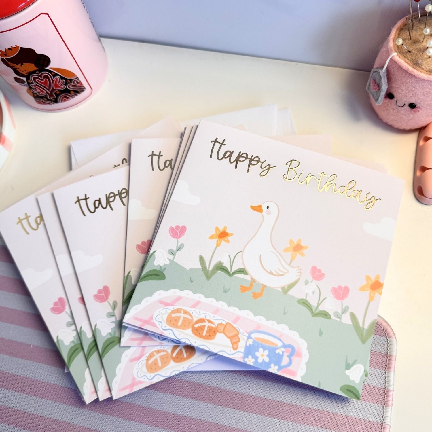 A stack of pastel-colored 'Happy Birthday' greeting cards featuring a cute white duck, yellow flowers, and a picnic scene, styled on a striped surface with pink accessories in the background.